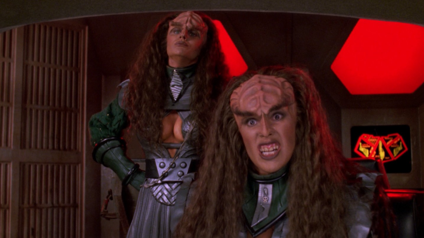 Two Klingon warriors in metallic armor stand in a red-lit sci‑fi set, one snarling toward the camera and showing teeth.