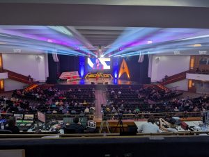 Auditorium stage with large video screen showing a speaker, surrounded by purple and blue lighting and an audience watching from rows of seats lighted by colorful beams.