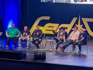 Panel discussion on a stage: six men seated in a row, some with drinks, beneath a large yellow logo and blue lighting; one man raises a hand as he speaks.