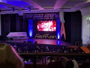 Stage at a sci‑fi convention hall with blue lighting, a large central screen showing bright graphics, banners on either side, and an Enterprise model on a pedestal; audience seated.
