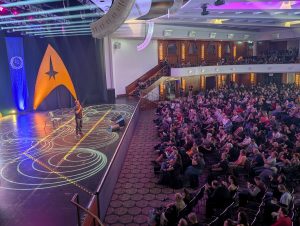 A performer on a lit stage beside a large orange triangular prop and a blue emblem banner, addressing a seated audience in a theater venue.